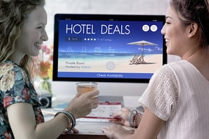 Dos mujeres reservan unas vacaciones en línea viendo ofertas de hotel en una pantalla de escritorio con imagen de playa.