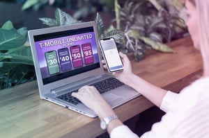 Persona compara planes ilimitados de T-Mobile en una laptop mientras sostiene un teléfono móvil.